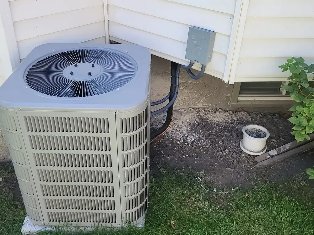 A central air conditioner beside a house with cream siding, its disconnect box on the wall above