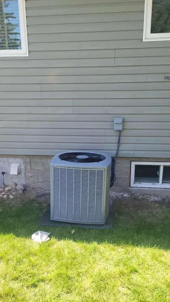 A central air conditioner on a pad in the lawn beside a house with grey siding