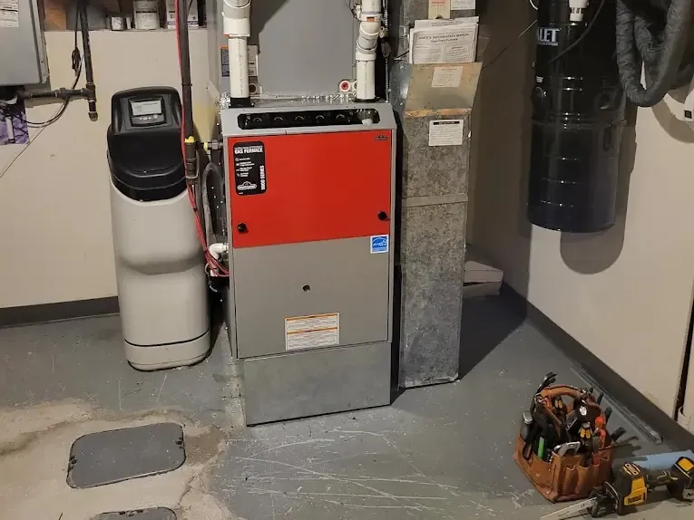 A furnace with a red front panel in a basement utility room, next to a water softener and a tool bag